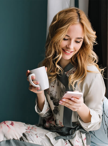 pretty-blonde-girl-sitting-window-sill-with-cup-coffee-tea-smartphone-hands-she-has-long-blonde-wavy-hair-smile-looking-her-phone-wearing-beautiful-silk-pajama